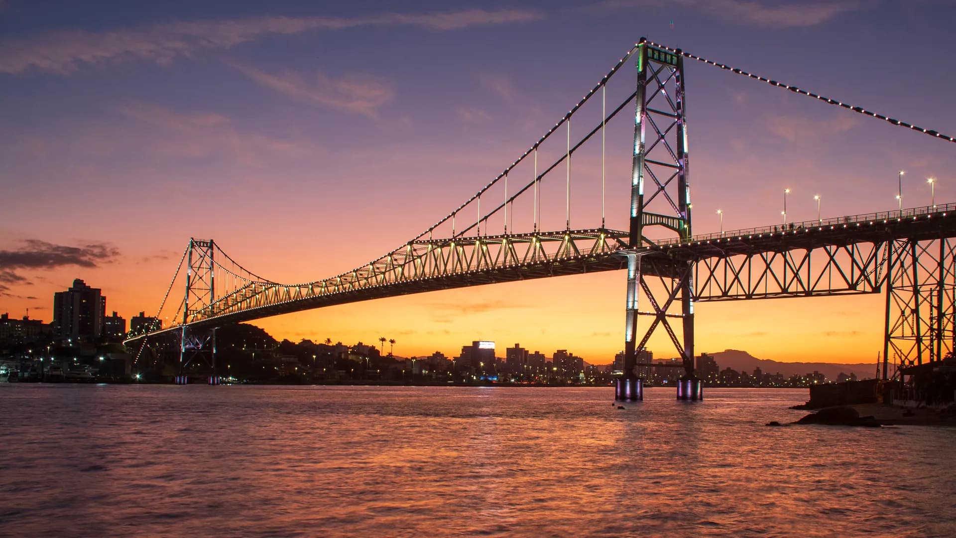 A Ponte Hercílio Luz iluminada ao entardecer, vista da baía de Florianópolis
